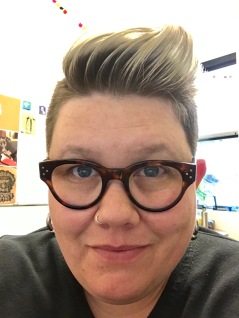full face shot of a caucasian butch female with a dark roots/silver tips pompadour looks directly into the camera. she has blue eyes, silver earrings, and a black v-neck sweater on.