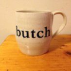 a handmade cream colored mug has the text "butch" on it in black serif lettering; the mug sits on a wood table in front of a cream wall